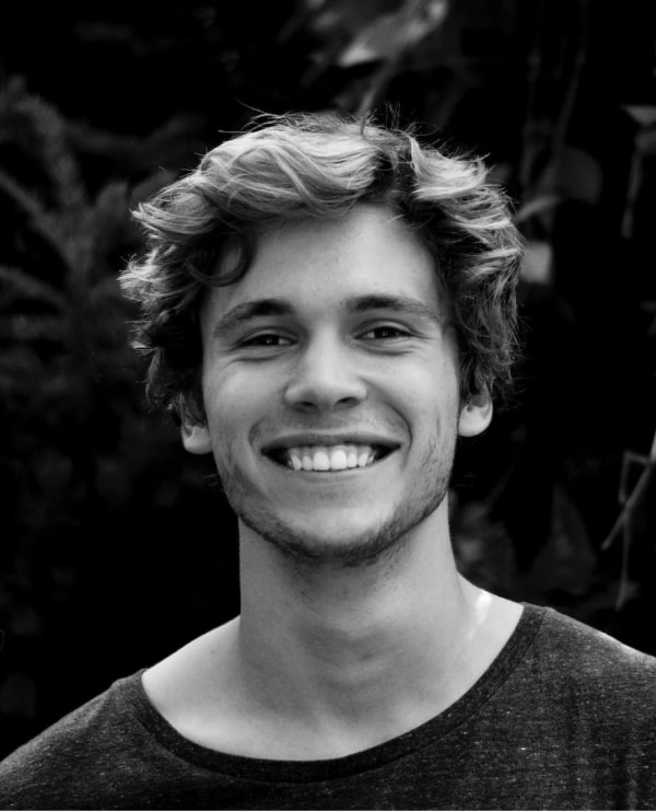 Cheerful young man smiling in black and white against vibrant greenery backdrop.