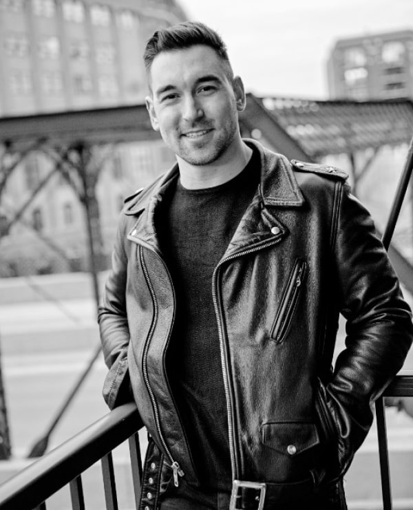 Stylish man in leather jacket smiles confidently against an urban industrial backdrop.