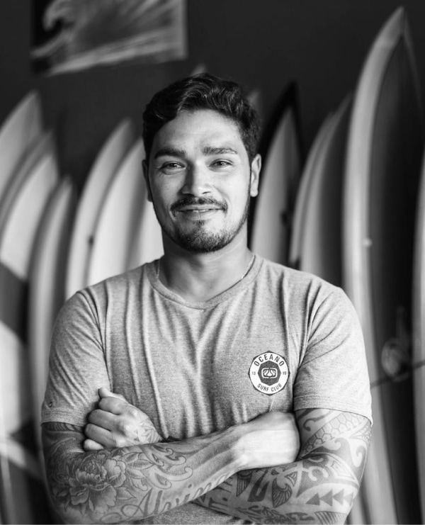 Confident young man with tattoos, smiling amidst surfboards, embodying the surfing lifestyle.