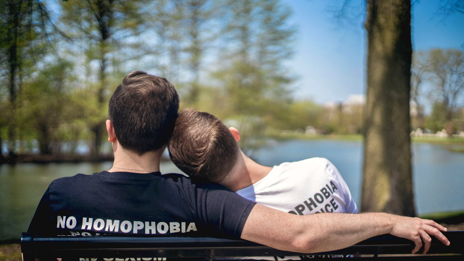 Dos hombres sentados en un banco de un parque junto a un lago, abrazando el amor con unidad y orgullo.