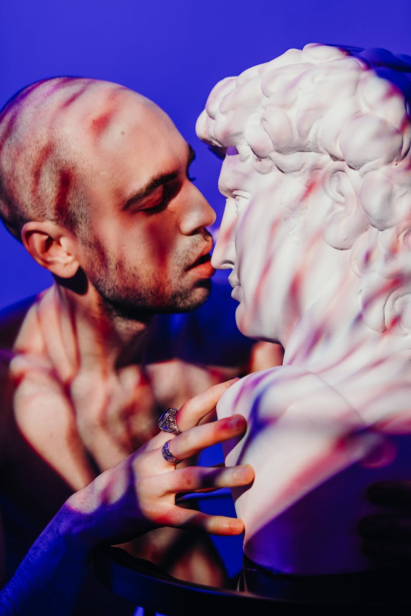 Un homme chauve et tatoué interagit intimement avec une sculpture classique sous une lumière colorée.