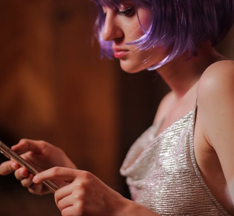 Young woman in violet wig and sparkly dress using a smartphone indoors.