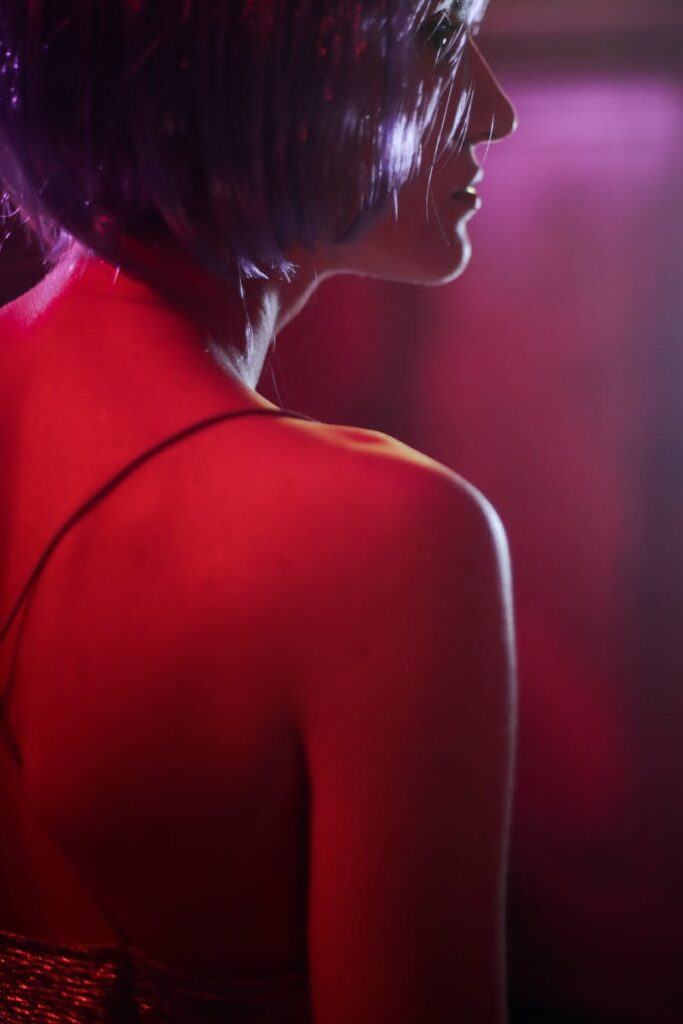 A woman in profile with a purple wig, illuminated by neon light, wearing a spaghetti strap dress.