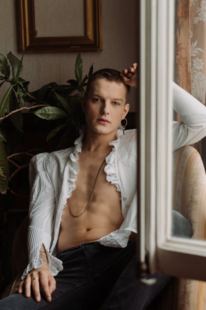 Hombre sin camisa con atuendo elegante sentado junto a la ventana con luz natural.