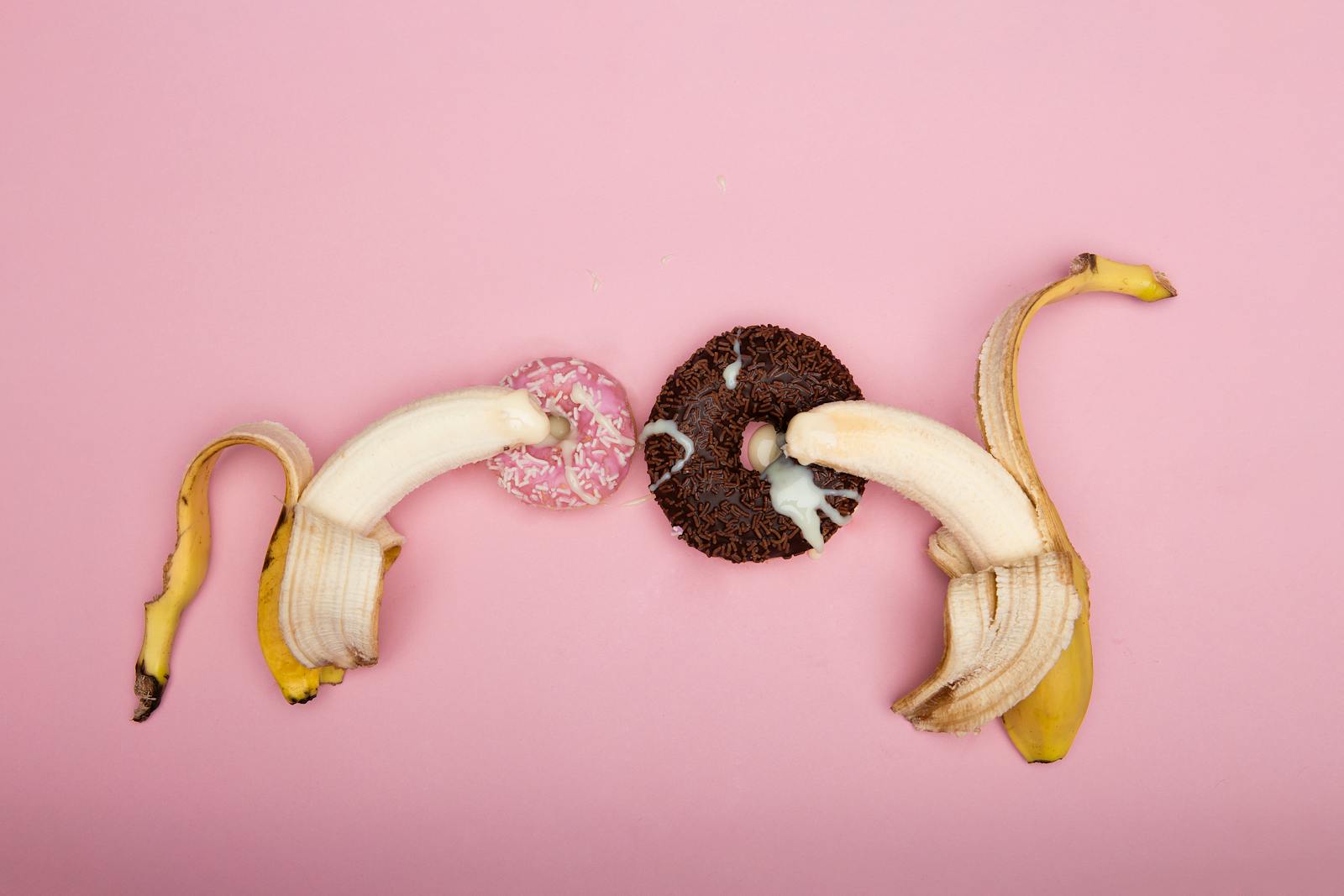 Speelse opstelling van gepelde bananen en donuts op een roze achtergrond, perfect voor conceptuele kunst.