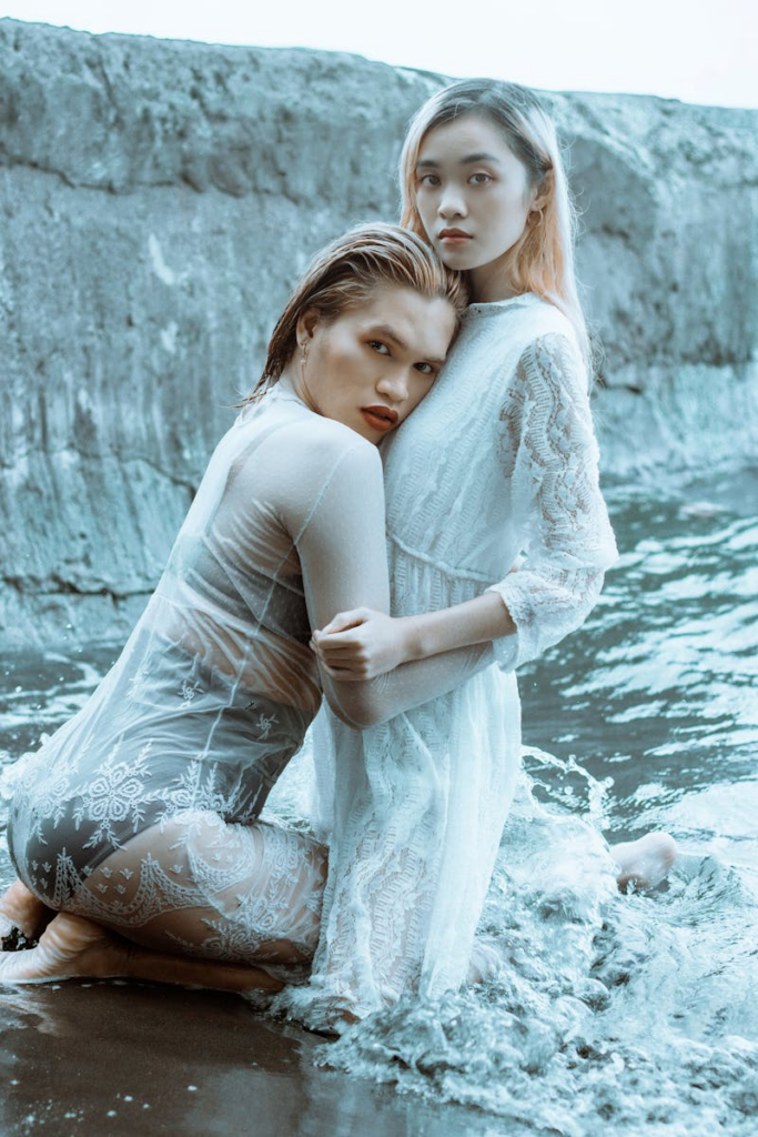 Side view of barefoot young woman hugging with Asian girlfriend in white elegant dress while spending time on seashore with boulder in daylight