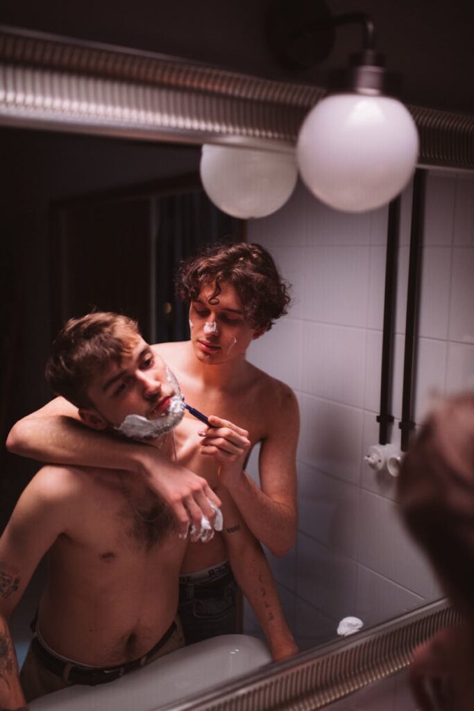 Two men sharing an intimate shaving moment in a bathroom mirror reflection.